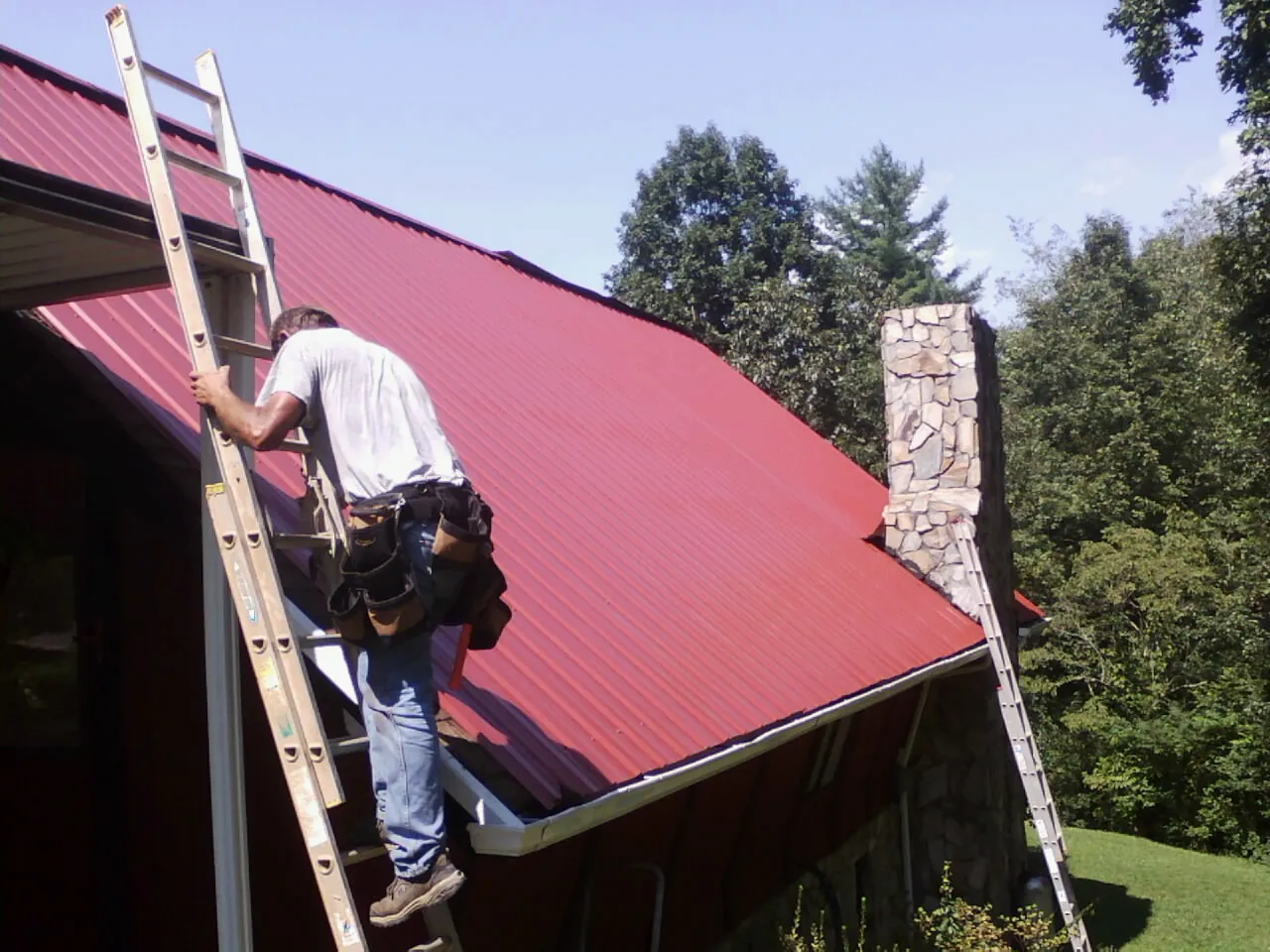 Roofer on a ladder beside a red metal roof and stone chimney — Soffit & Fascia Repair by Roofing Brothers in Three Rivers